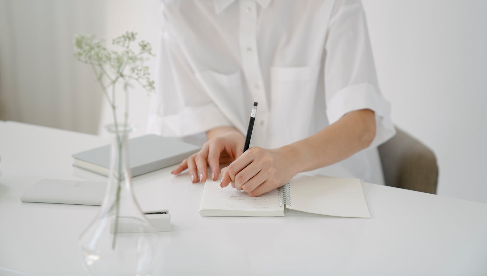 Photo by John Diez A woman writing in a notebook at a minimalist desk with stationery items and a vase.